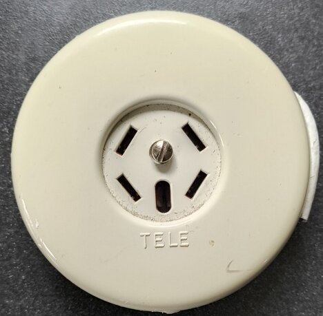 Rund vit äldre telejack i plast monterad på en grå vägg. Fyra anslutningsöppningar och text "TELE" skrivet under.