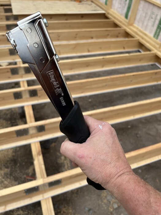 En hand håller en klammerhammare med märkningen Rapid R34 mot bakgrund av en träkonstruktion.