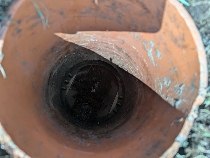 En öppen rödbrun avloppsrörstopp syns nedifrån, inuti syns smuts och rörkanter.