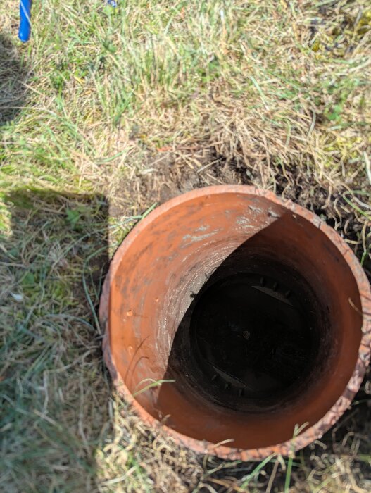 Öppen brunn med brunt rör i marken, omgiven av gräs.
