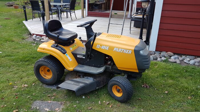 Gul Partner åkgräsklippare modell 12-105E vid en träveranda, med stengång och rödmålad byggnad i bakgrunden.