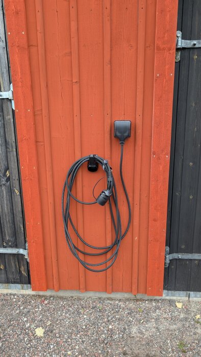 Vägg med en elektrisk dosa och en upphängd kabel på en orange träfasad, nära en svart dörr.