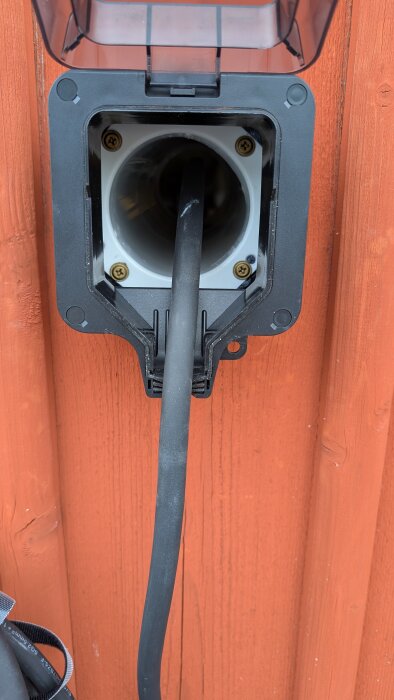 En kabel dras genom en anpassad ute dosa med en gummitätning, monterad på en trävägg för inomhusladdning.