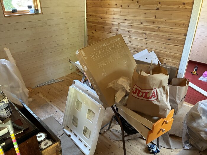 Rum under renovering med trägolv och väggar, kartonger, plastpåsar och verktyg från Jula liggande på golvet och möbler.