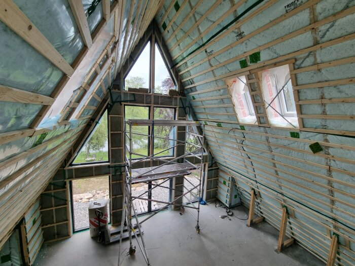Ett rum under byggnation med triangulära fönster, isolering på väggarna och ställningar uppställda för arbete.