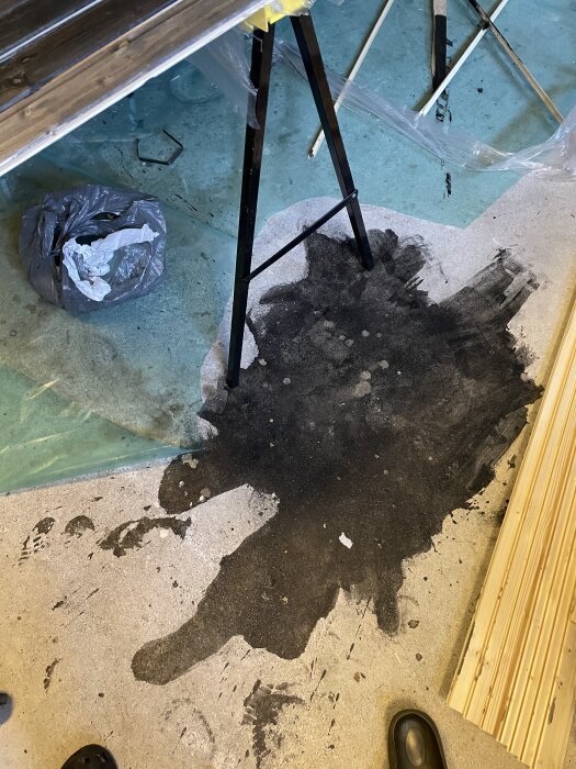 Svart vax spillt på garagegolv bredvid målade panelbrädor och plastpåse.