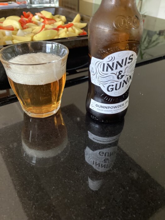 Flaska med Innis & Gunn-öl och glas på ett svart bord, i bakgrunden ses råstekt potatis och paprika på en plåt.