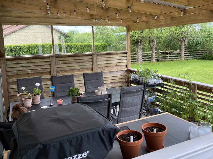 Övertäckt altan med sittplats, blommor och fönsterkassetter. Grön gräsmatta med träd i bakgrunden.