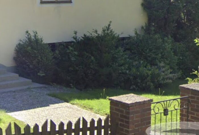 Husfasad med buskage och växtlighet nära trottoar och staket.