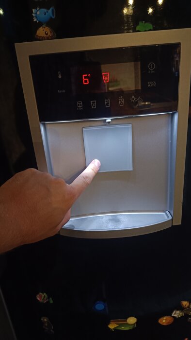 Kylskåp med en hand som pekar på dispensern, displayen visar 6 grader och ikoner för vatten och kyldryck.