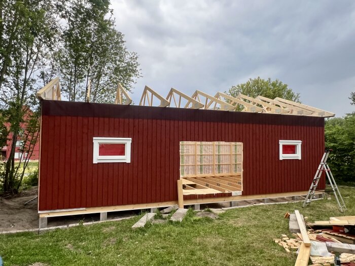 Rött hus med färdiga takstolar, två fönster och en stege på sidan.