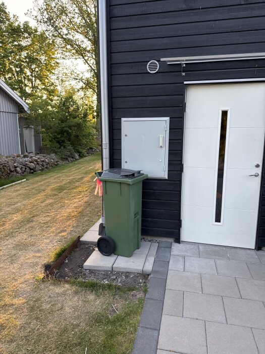 Grön soptunna på plattor vid svart husvägg, med gräsmatta bredvid och stenar i bakgrunden.