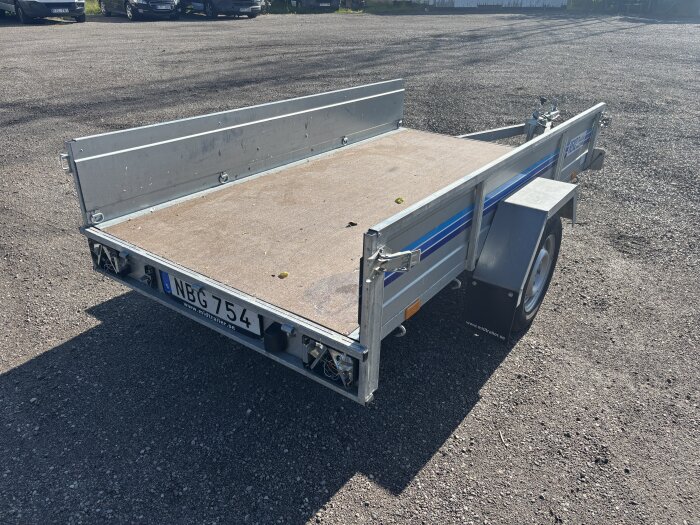 En släpvagn med registreringsskylt NBG 754 på en grusparkering. Saknar delar av bakre lampglas och har lämmar fram och bak samt träbotten.