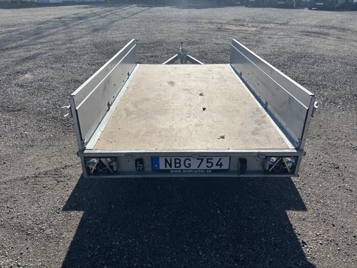 Släpvagn med öppen lastplattform, saknar baklampor och har registreringsskylten "NBG 754". Står på grusplan.