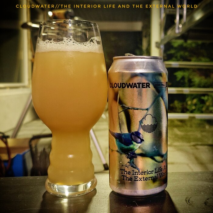 Ett glas med disig öl bredvid en Cloudwater-burk med etiketten "The Interior Life And The External World" på ett bord utomhus.