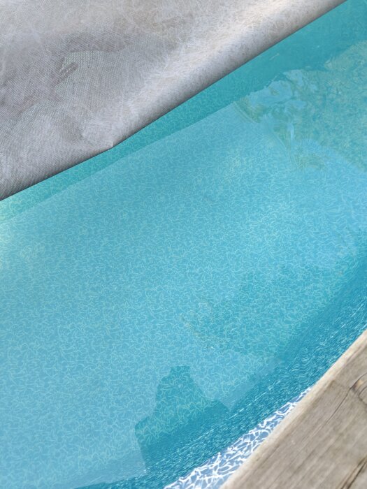 En pool med klarblått vatten och ett stenmönstrat bottenmönster, delvis täckt av ett grått tyg eller skynke.