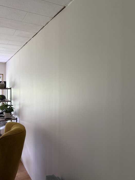 Ojämna spackelskarvar syns på en vägg med blek målning. Takets skarv vid väggen är också markerad. Möbel och hylla i bakgrunden.