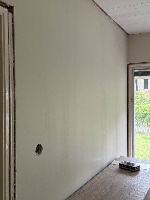 Vägg i vardagsrum med synliga spackelskarvar och ojämn målningsyta, med bord framför och utsikt mot trädgård genom fönstret.