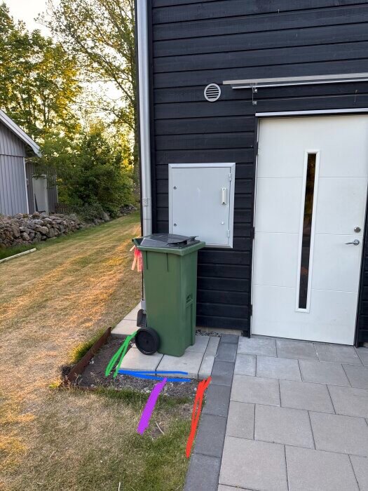 Grön soptunna vid svart fasad, bredvid vit dörr. Färgade linjer på marken visar möjliga kabeldragningar. Bakgrund med gräsmatta och stengång.