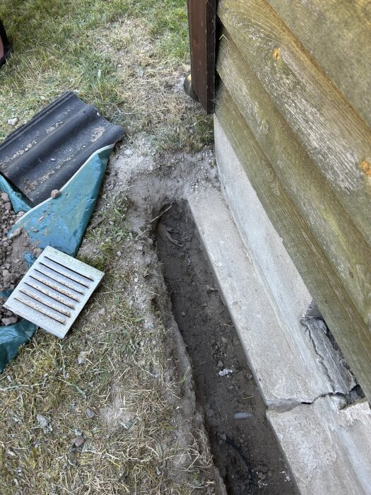 Ränna grävd längs husvägg med jord och gräs runt. En bit markduk och ett galler ligger på marken bredvid, förberett för fyllning och lutning.
