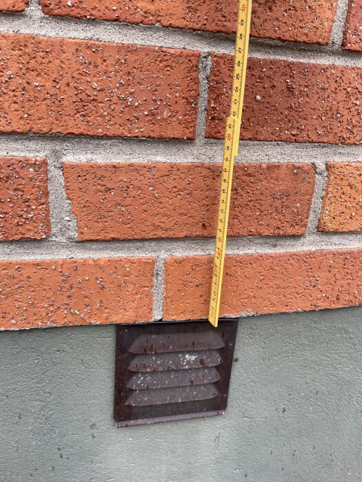 Tegelvägg med murbruk mellan plattorna, ett lodrätt måttband som visar 20 cm och en kvadratisk, ventilerad metallplåt i nederkant.