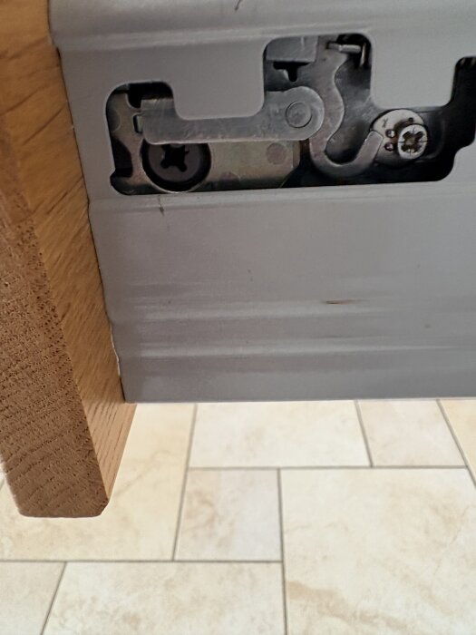 Lådmekanism från Ballingslöv med skruv och kopplingsdetaljer, del av lådfront synlig i trä, mot ljus kakelbakgrund.