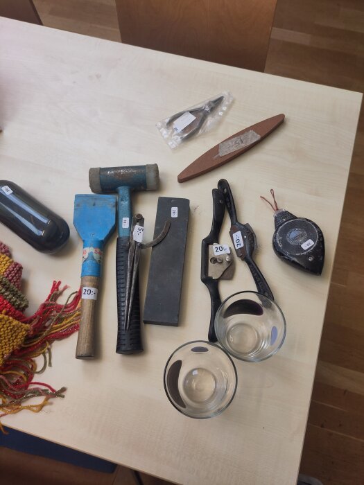 Verktyg och föremål på ett bord, inklusive hammare, tång, måttband, slipsten, två glas och färgglada tyger, alla märkta med prisetiketter.
