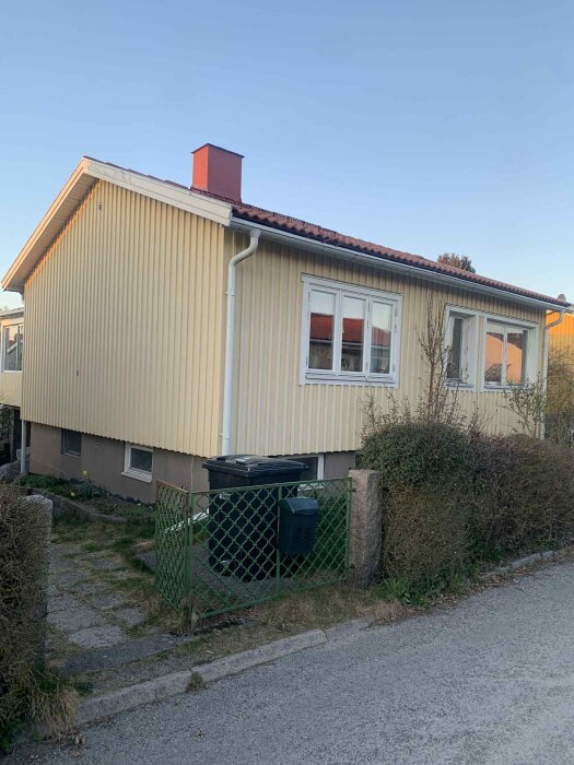 Gult hus med rött tak och vit fönsterkarm, bredvid grön grind och soptunna vid trottoaren, buskar runtomkring.