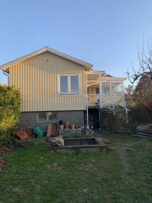 Gul villa med anslutande uterum och trappor, gräsmatta fram och trädgårdsredskap längs husets källarvägg.