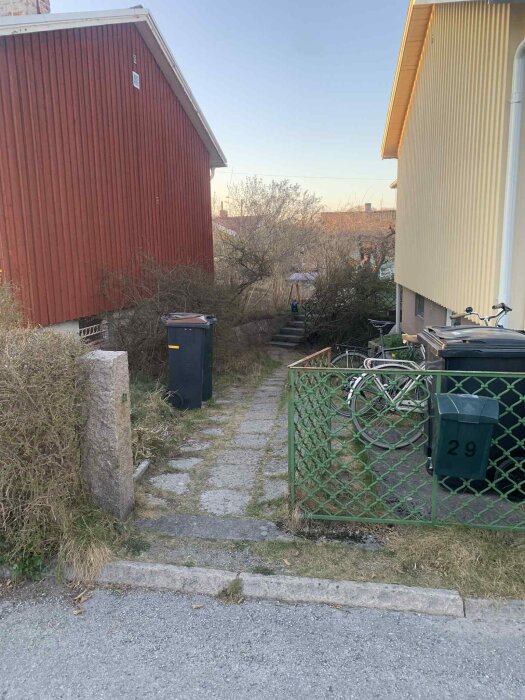 Gångväg mellan röd och gul husfasad med grön grind, stenmurar och cykelparkering. Grönska och soptunnor syns längs vägen.