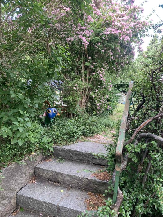 Stentrappa omgiven av grönska och buskar med rosa blommor, en dekoration med fågelmotiv syns bland växterna.