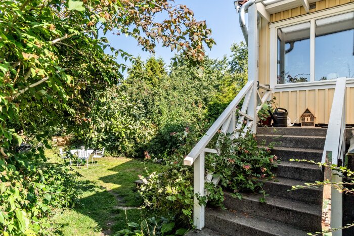 Trappa med vita räcken leder upp till ett uterum på ett gult hus, omgiven av lummig trädgård och gräs.