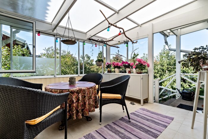 Inglasat uterum med svart rottingmöbler, blommor och dekorationer, med utsikt över grönska genom glasfönster och ett ljust tak ovanför.