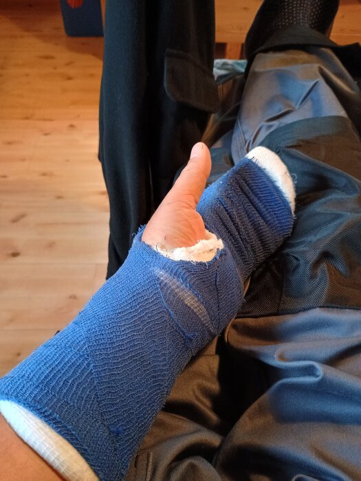Blå gipsad vänsterhand vilande på mörka byxor, inomhus med ljus trägolv i bakgrunden.