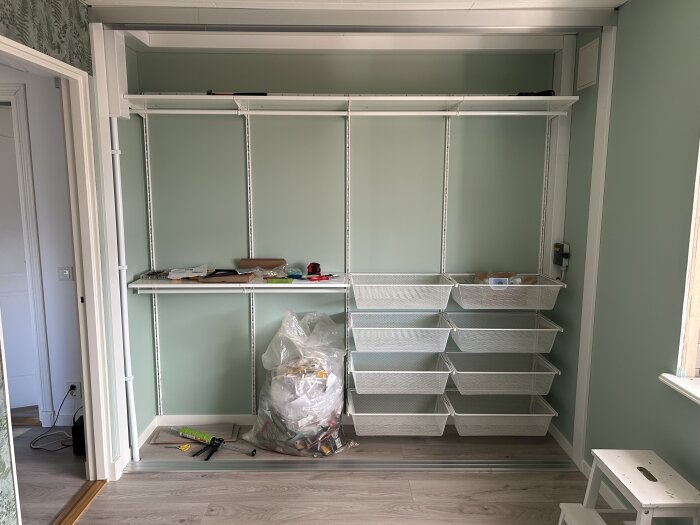 En oavslutad garderob med hyllor och gallerlådor, verktyg och material på golvet framför, del av pågående renoveringsprojekt.