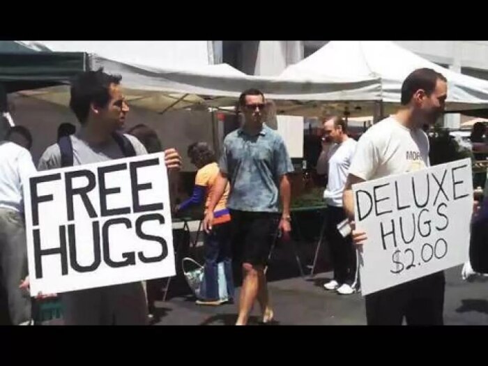 Två personer står utomhus, en håller en skylt med "Free Hugs" och den andra med "Deluxe Hugs $2.00". Flera människor syns i bakgrunden.