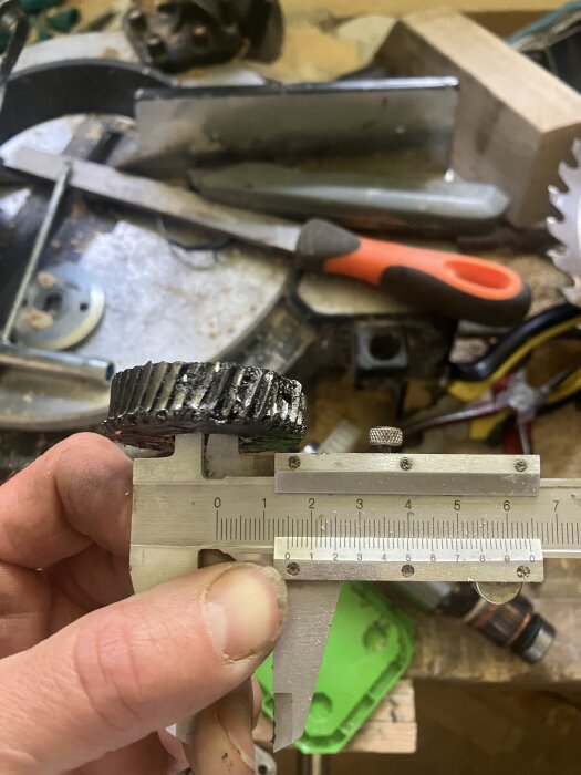 Hand holding a caliper measuring a worn, greasy gear on a cluttered workbench with tools in the background.