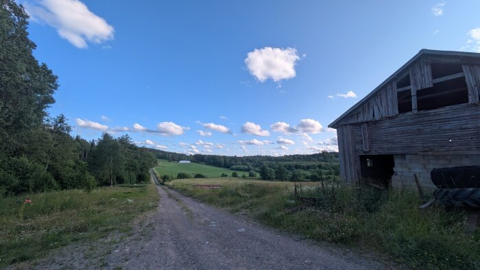 Landsväg genom grönskande ängar med en gammal lada på höger sida under en klarblå himmel med vita moln.