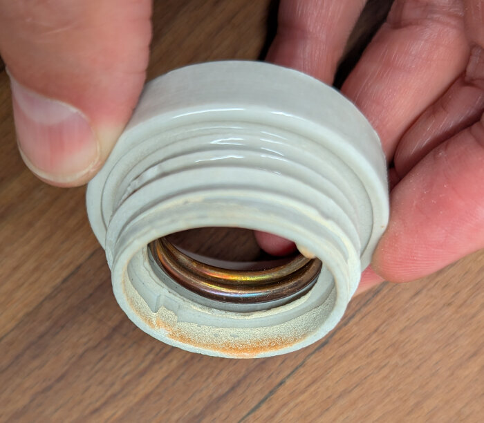 Närbild av en hand som håller en porslinslamphållare med metallbleck, visar slitage och rost på botten.