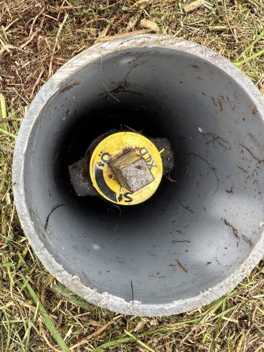 Gul ventil i grå ett-rörs-tub ovan jord, delvis omgiven av gräs och jord, visar en stängningsanvisning.