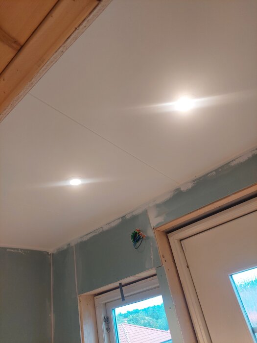Nyrenoverat rum med nymålat tak och installerade taklampor över ett fönster och dörr, med synliga kablar i väggen.