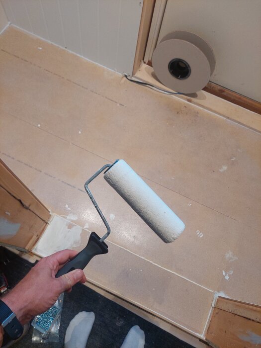 Hand håller en roller ovanför ett nyligen primer-behandlat golv, med en kabelrulle bredvid.