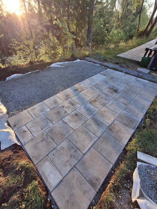 Plattor lagda i ett rutmönster på marken nära ett skogsområde, med grus och trätrall i bakgrunden.