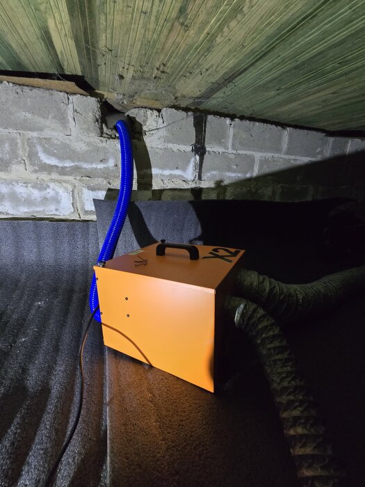 En orange Drybox-enhet är igång med blå och svarta rör anslutna, placerad vid en tegelvägg i ett utrymme.