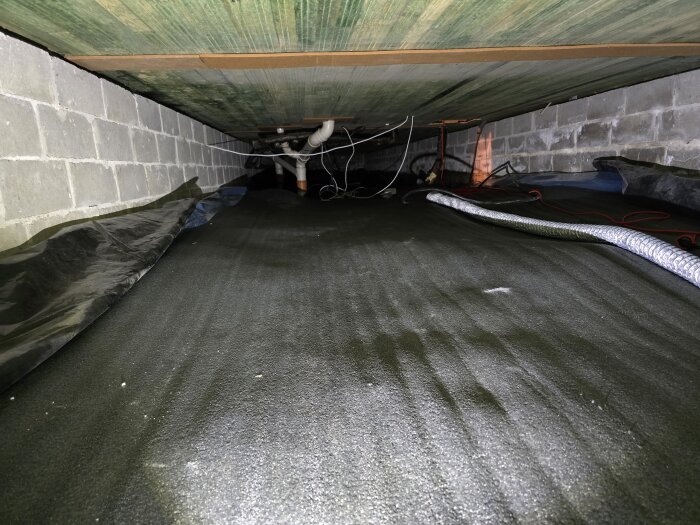 Krypgrund med tejpad plast på golv och väggar, ventilationsrör samt en rörelse i rör och sladdar längs tak och väggar.