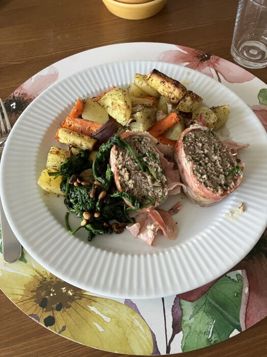 Baconlindad köttfärslimpa, ugnsrostade grönsaker, sauterad spenat med pinjenötter på en tallrik med blommigt underlägg.