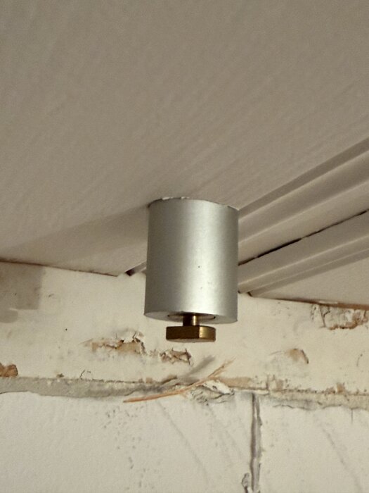 Silverfärgad cylinder hängande från innertaket i ett hus, med en skruv eller bult undertill; väggen visar tecken på slitage.