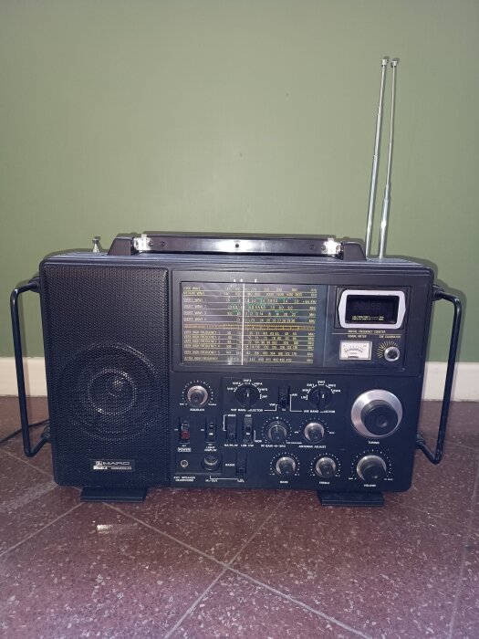 Gammaldags radioapparat med flera rattar och antenn synlig mot en grön bakgrund.