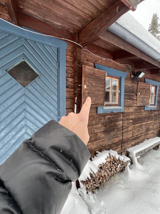 Hand pekar på stuga med blå dörr och timmerväggar i snötäckt omgivning, diskussion om färgval och behandling av träet i inlägget.