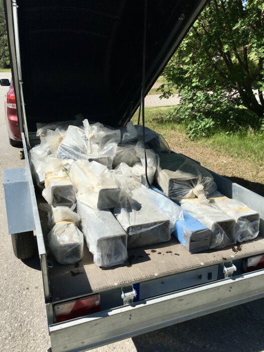 Släpvagn fylld med plastförpackade rör, delvis inlindade, för transport till deponi, står parkerad vid en väg.